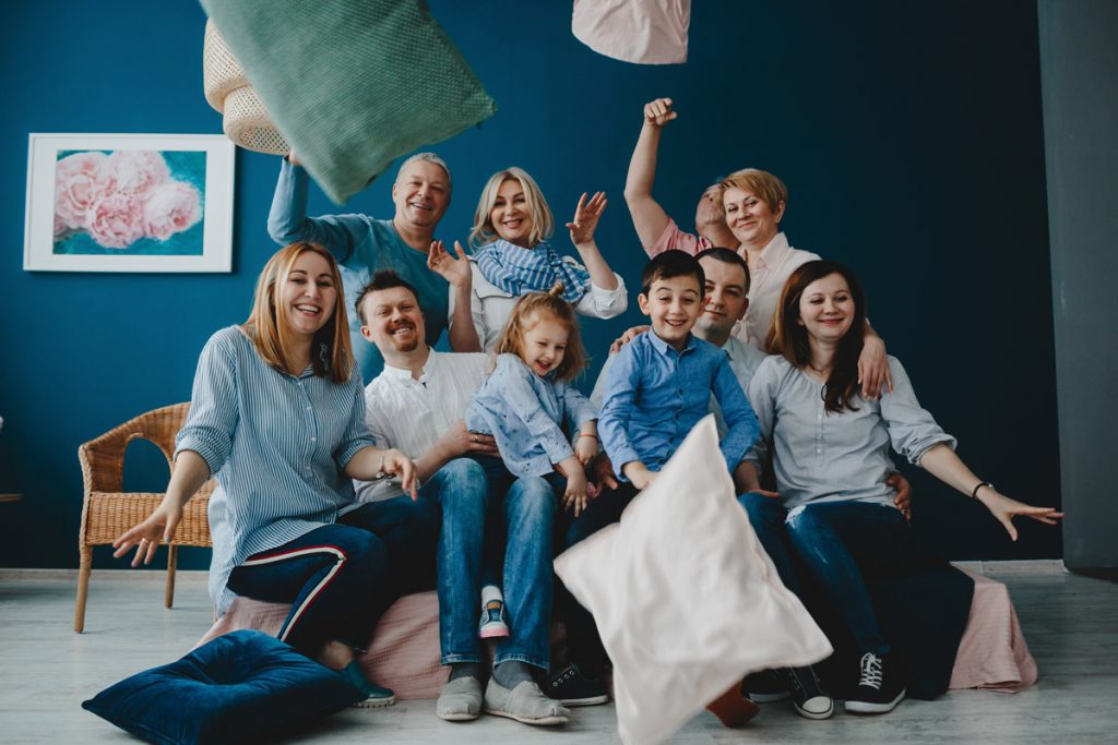 Grandparents, parents and their little children sit together on the bed in a blue room and fight pillows.